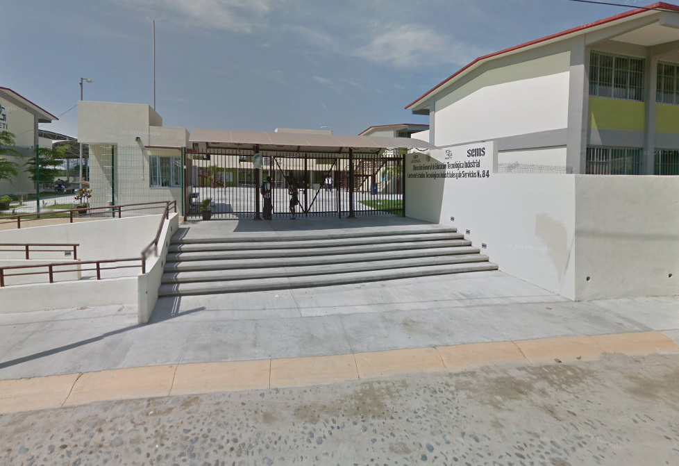 Fotografía en la que se muestra la fachada del plantel conformada por una reja roja y a su lado derecho hay un muro de color blanco, que en letras negras dice el nombre del plantel. Frente a la reja hay una escalera y una rampa que dan acceso a las instalaciones.